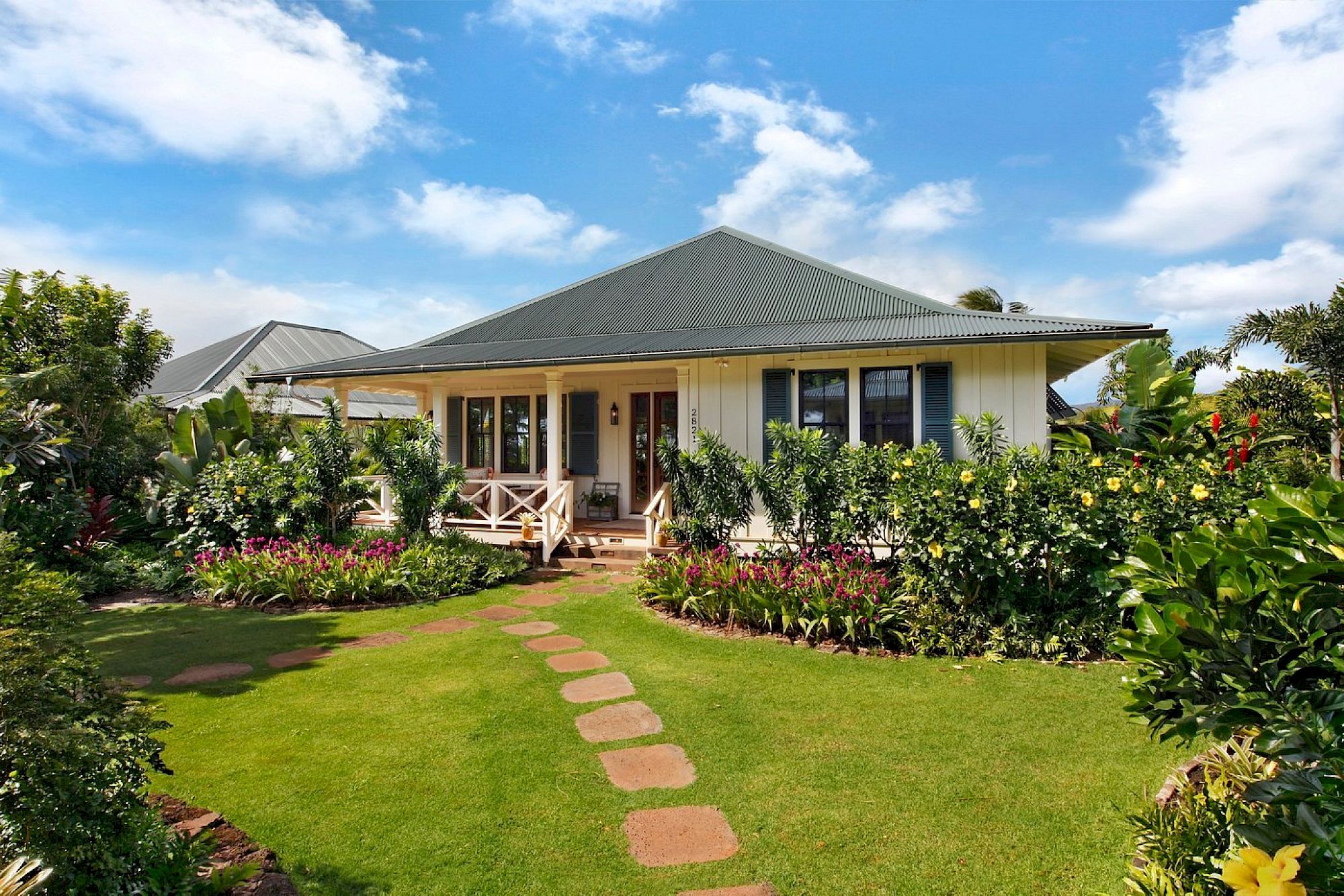 A sunny tropical bungalow with a wide veranda, surrounded by lush gardens and a stone walkway under blue skies.