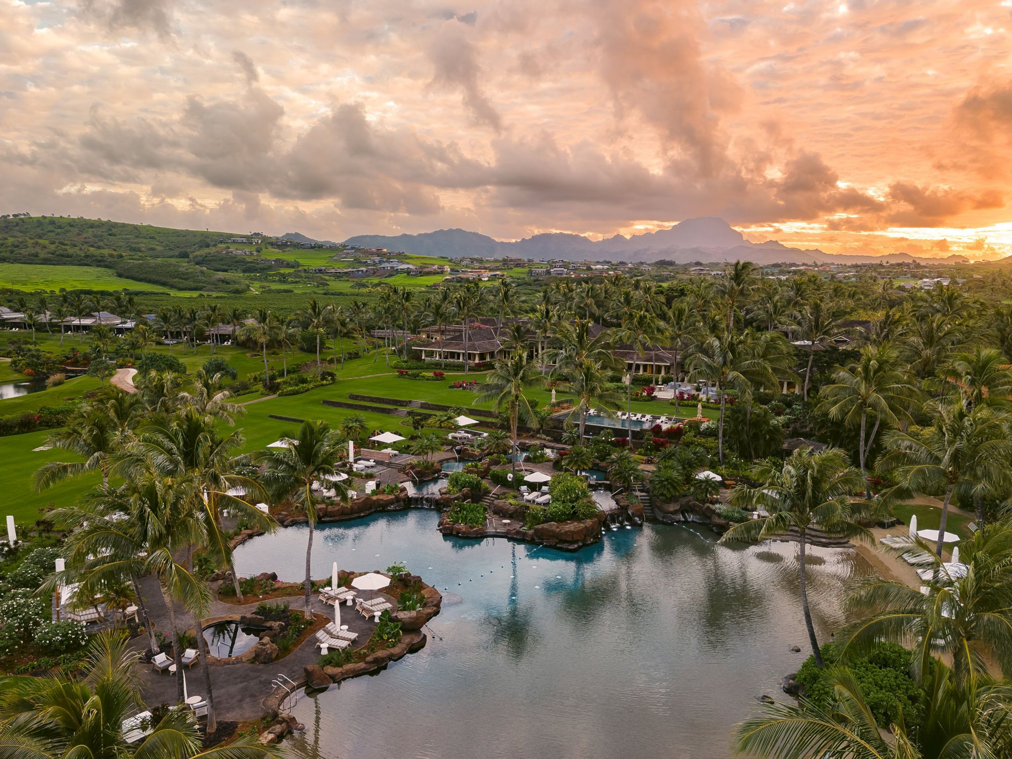 A tropical resort with palm trees, green lawns, and a lagoon-like pool by a sunset over rolling hills and mountains.