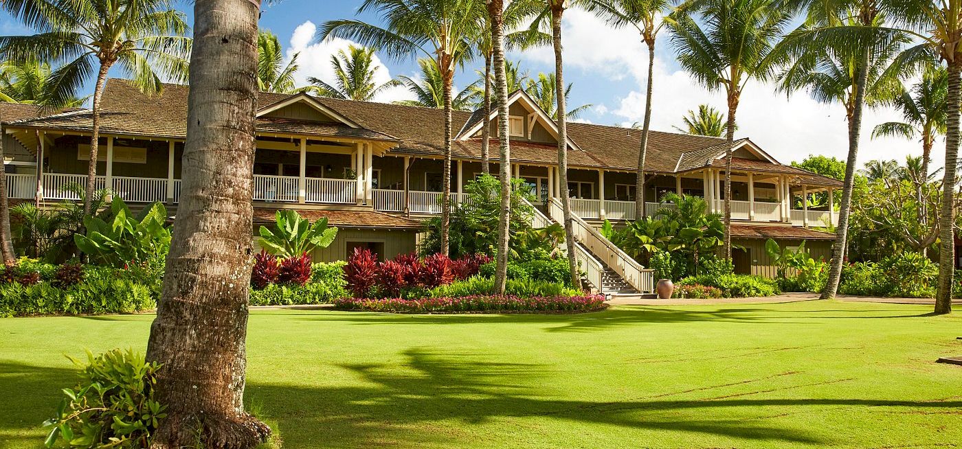 A tropical resort scene with tall palm trees, manicured green lawn, and a long balcony building under a blue sky.