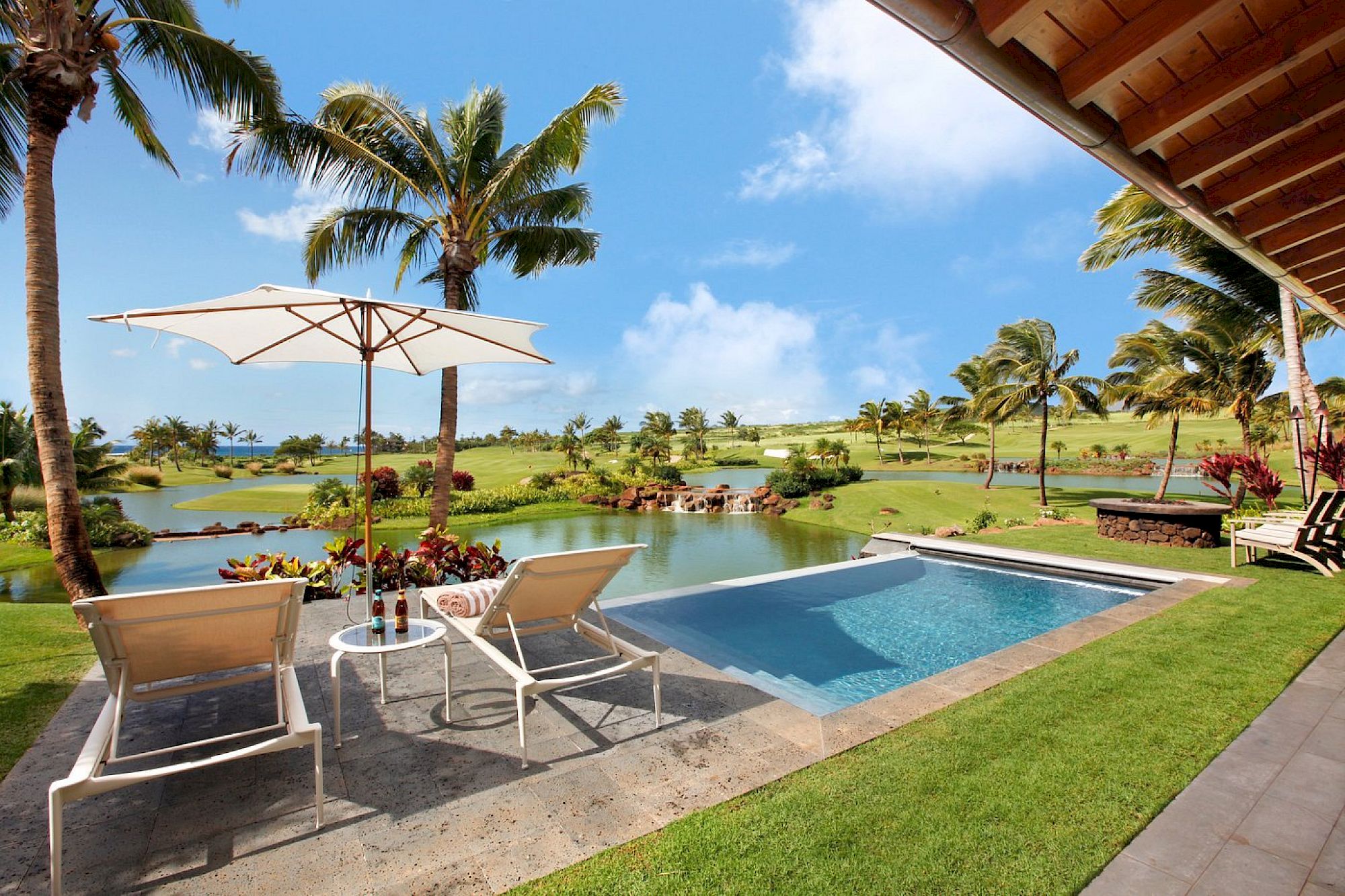 A sunny tropical resort patio with a pool, lounge chairs, a white umbrella, palm trees, and a scenic pond in view.