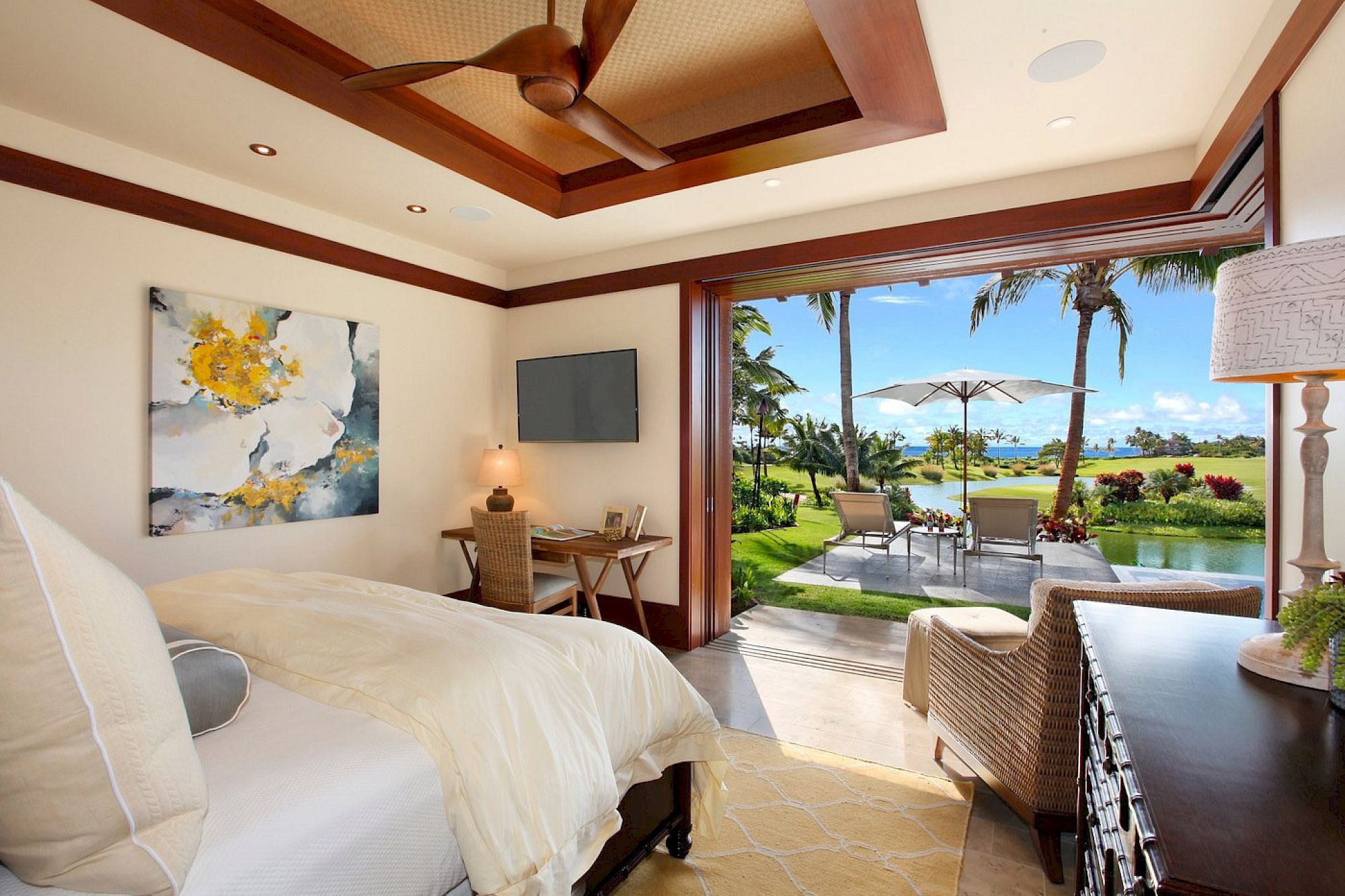 A luxurious bedroom with a ceiling fan, framed art, a TV, and open doors showing a patio with umbrellas, chairs, and a tropical ocean view.