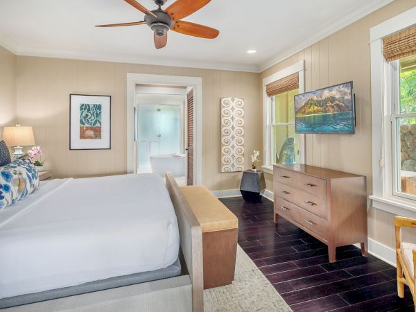 Bright bedroom with a ceiling fan, large bed, dresser, flat-screen TV, big windows, and beachy decor.