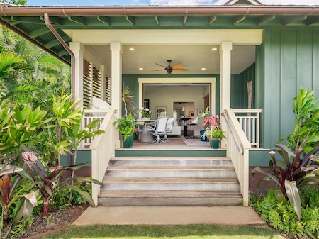 A tropical front porch with green siding, steps leading to a covered veranda, and lush plants with a ceiling fan.
