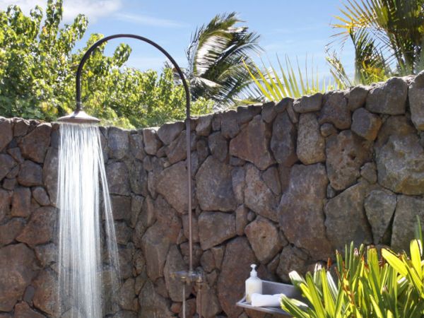 An outdoor garden fountain spills water from a spout into a stone wall, with palm trees and green plants nearby.