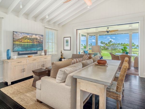 Bright tropical living room with a large TV, beige sectional, dining table, and open patio doors overlooking green scenery.