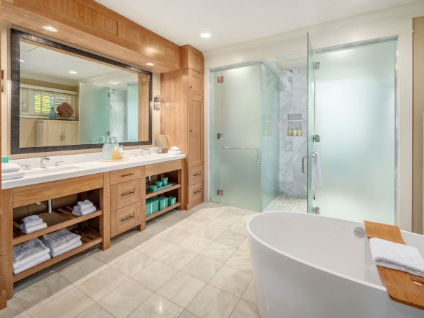 A bright modern bathroom with double vanity, mirror, glass shower, and a white soaking tub with folded towels in view.