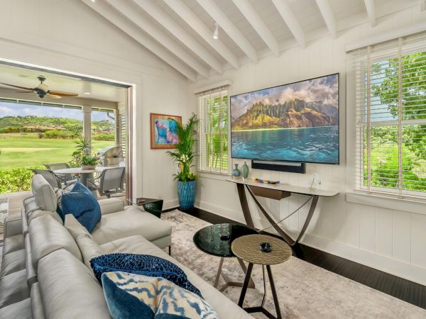 A modern living room with a large wall TV, comfy white sectional sofa, coffee tables, and bright windows overlooking green hills.