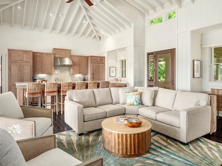 A bright living room with beige sofas, woven chairs, a round wood coffee table, patterned rug, and a visible kitchen with stools.