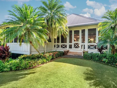 A tropical resort home with a veranda, framed by lush palm trees, colorful gardens, and a green lawn in front.