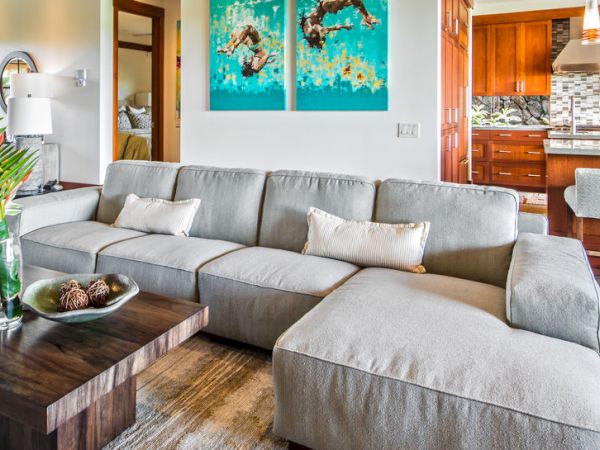 A modern living room with a gray sectional, wooden coffee table, and an adjoining wood-paneled kitchen with stools and pendant lights.