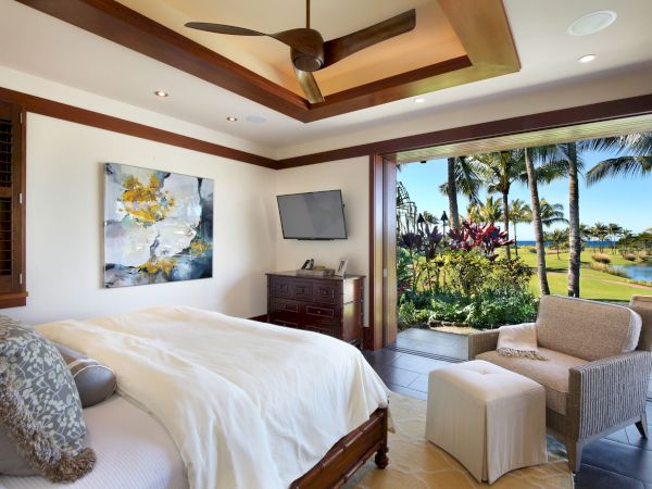 A luxury bedroom with a ceiling fan, TV and dresser, opening to a sunny patio with palm trees, lawn, and ocean view.