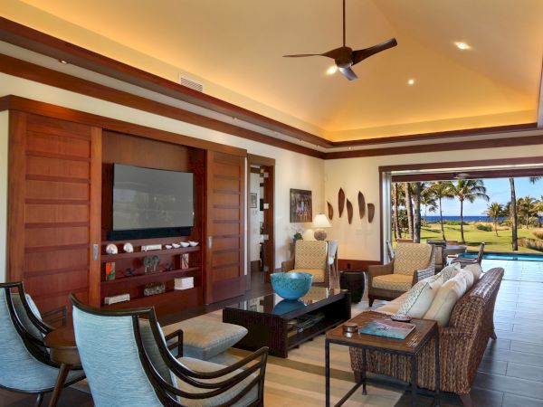Sunlit living room with wood wall, flat-screen TV, ceiling fan, and patio view of palm trees and the ocean.