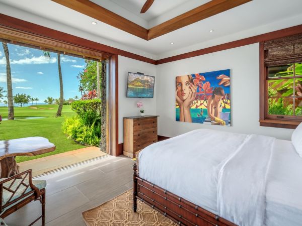 Sunlit tropical bedroom with a large bed, framed art, and open doors revealing a green lawn by the ocean.