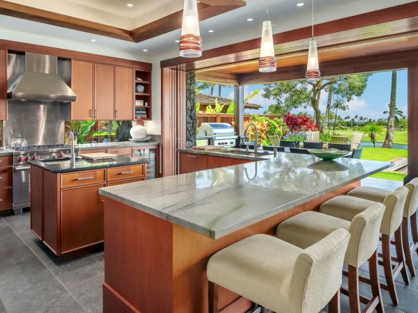 Luxurious outdoor kitchen with a stainless grill, granite island, bar stools, and a view of a tropical garden and lawn.