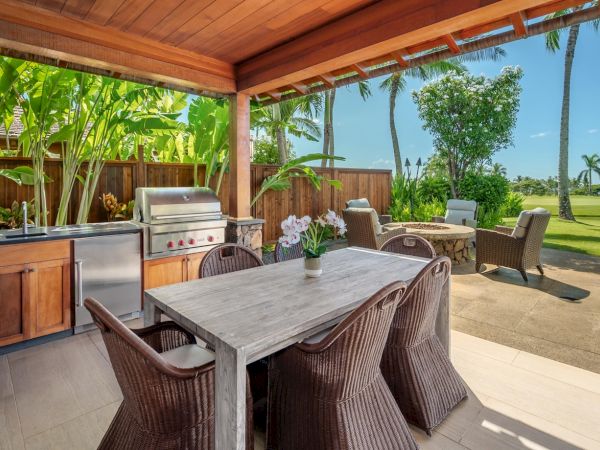Tropical outdoor patio with a stone wall, covered dining table, wicker chairs, and a stainless grill beside a landscaped yard.
