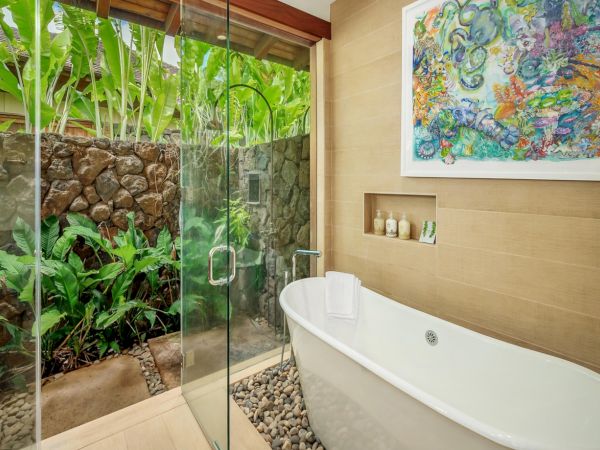 A modern bathroom with a glass shower, lush green garden foliage, a stone wall, a white soaking tub, and colorful artwork on beige tiles.