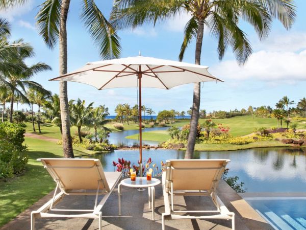 Two lounge chairs and a white patio umbrella overlook a tropical garden, palm trees, and a lake beside a golf course.