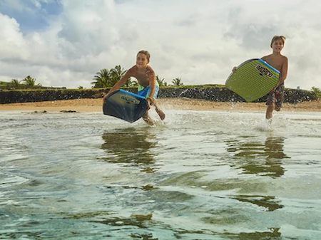 Two kids splash across a beach with surfboards, captured mid-jump on a cloudy shore.