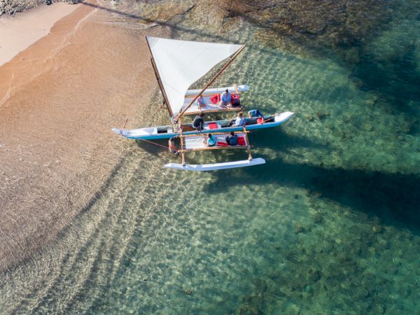Aerial view of two people on a sailboat with a beach and clear blue water, sailing near shore.