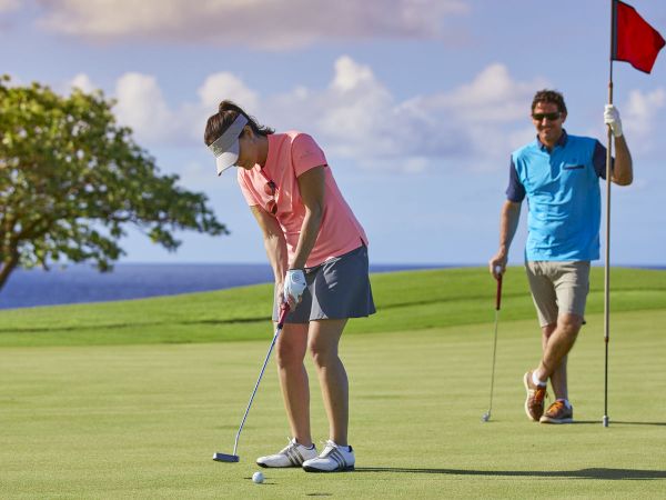 A golfer in pink lines up a putt on a sunny green while another player stands nearby with a flag.