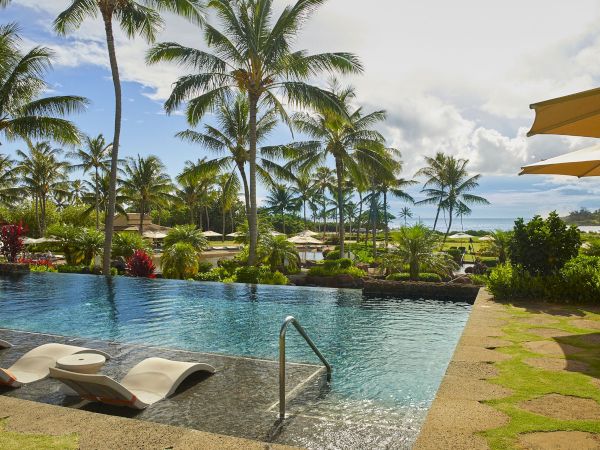 Tropical resort pool with palm trees, sun loungers, and yellow umbrellas overlooking the ocean under cloudy skies.