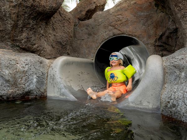 A toddler in a neon float rides into view from a water slide tunnel, wearing goggles and smiling.