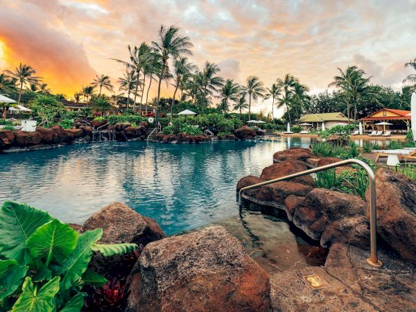 A tropical resort scene with a calm infinity-style pool, palm trees, sun umbrellas, and a warm sunset sky.