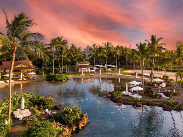 A tropical resort at sunset with palm trees, an outdoor pool, and white cabanas beside calm water.