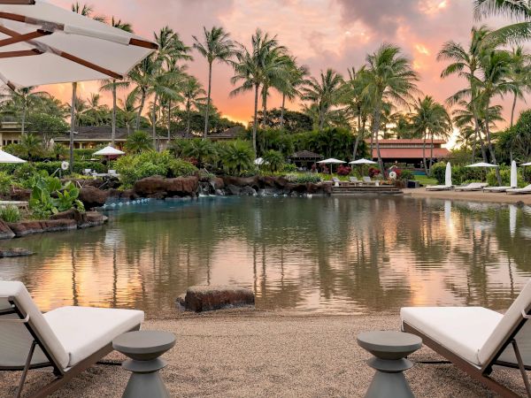 A tranquil resort pool at sunset with palm trees, lush gardens, sun loungers, umbrellas, and a calm reflecting waterline.