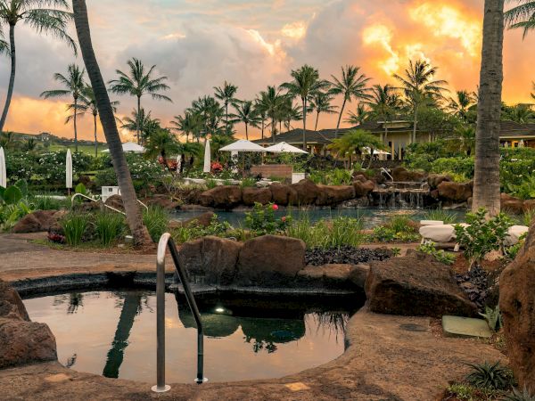 Tropical resort scene with palm trees, a serene pool and waterfall, and sunset clouds glowing over the gardens.
