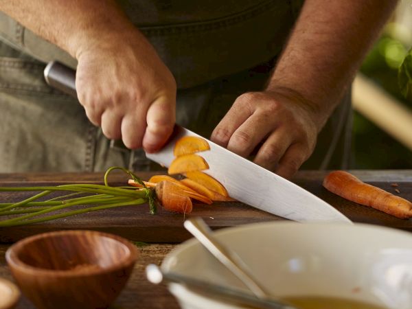 A person chops carrots on a wooden cutting board with a knife outdoors, with herbs and ingredients nearby.