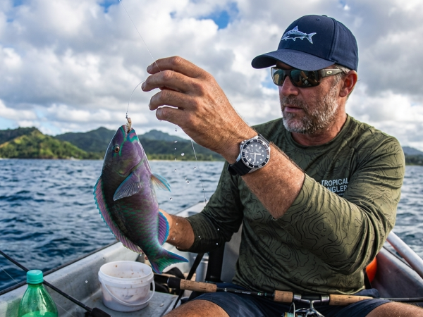 A man on a boat over choppy water holds up a colorful caught fish while fishing with a line and hook, in scenic mountains.