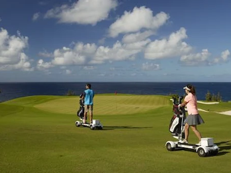 Two golfers ride motorized scooters across a seaside green course toward the holes under a bright blue sky.