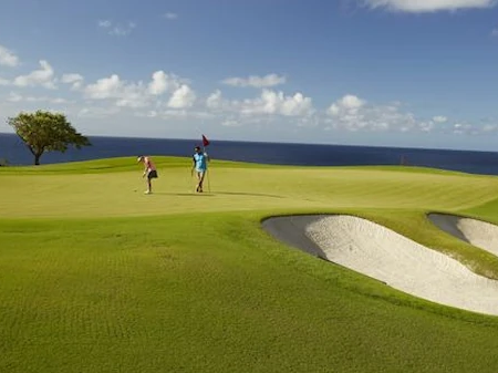 A golfer and companion walk along a sunny ocean-side fairway toward a sand bunker on a manicured golf course.