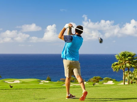 A golfer in blue swings a club on a sunny seaside green, with the ocean and palm trees in the background.