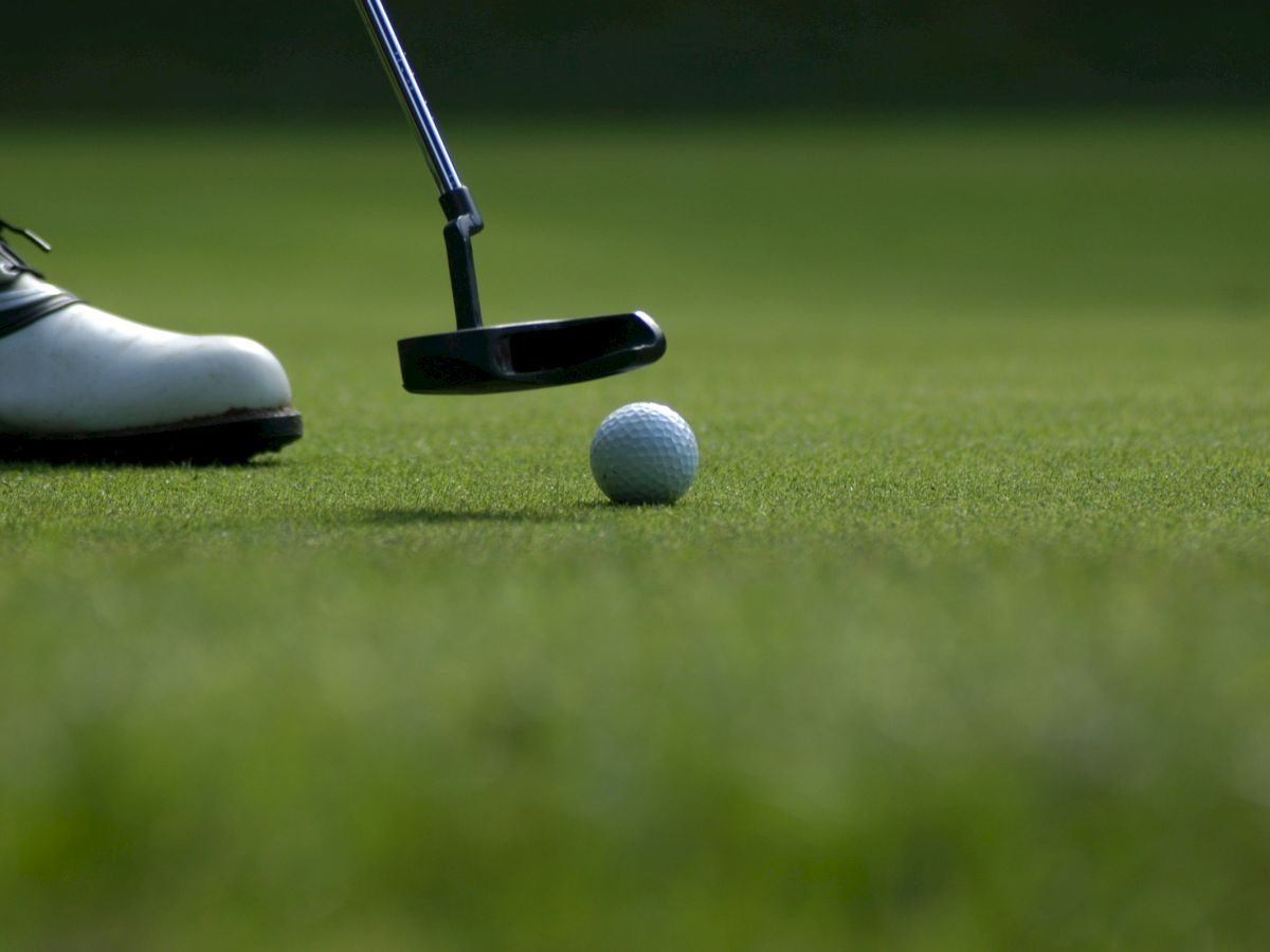 A silver golf ball rests on the green grass near a putter head, as if lining up a short putt on the course.