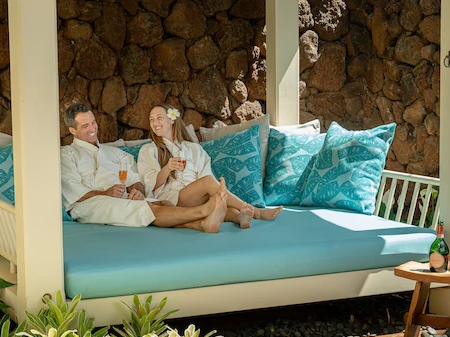 A couple lounges on a covered outdoor daybed, sipping drinks and relaxing, surrounded by pillows and greenery.