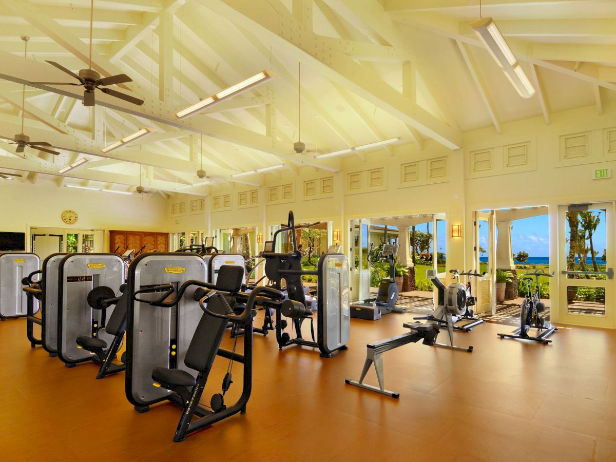 A bright gym filled with cardio and exercise machines, ceiling fans, and open windows showing the ocean view.