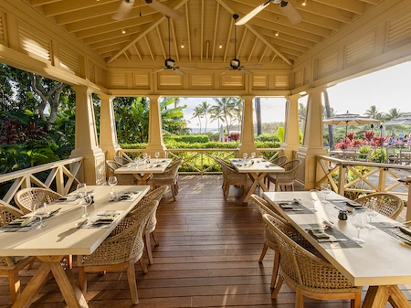 A bright open-air restaurant pavilion with wooden tables and chairs, surrounded by greenery and overlooking a tropical garden and water view.