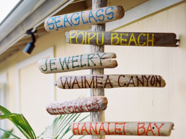 A weathered signpost at a beach listing destinations like &ldquo;Sealglass,&rdquo; &ldquo;Poipu Beach,&rdquo; &ldquo;JeWelry,&rdquo; &ldquo;Waimea Canyon,&rdquo; and &ldquo;Hanalei Bay.&rdquo;