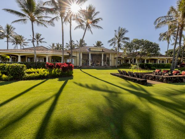A sunny tropical resort: manicured green lawn, palm trees, colorful flowerbeds, and a long hotel patio with chairs.