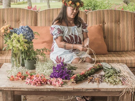 A young girl wearing a flower crown sits on a sofa while bundles of colorful flowers and greenery lie on a table in front of her.