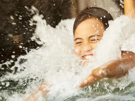 A young person smiles as waves splash around them, playing in churning water outdoors.