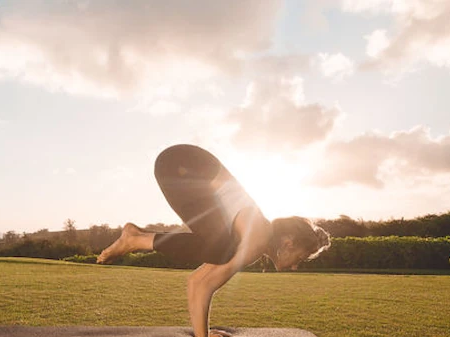 A person stretches in a yoga pose on a grassy field at sunset, with exercise equipment nearby.