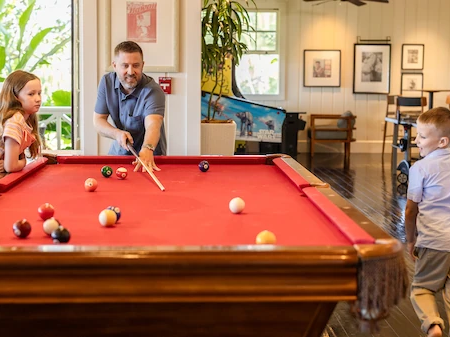 A family plays pool at a red billiards table in a bright room with framed pictures and plants.