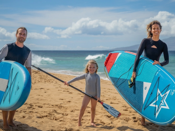 A family on a sunny beach holds surf-style inflatable paddleboards, smiling by the ocean under a cloudy sky.