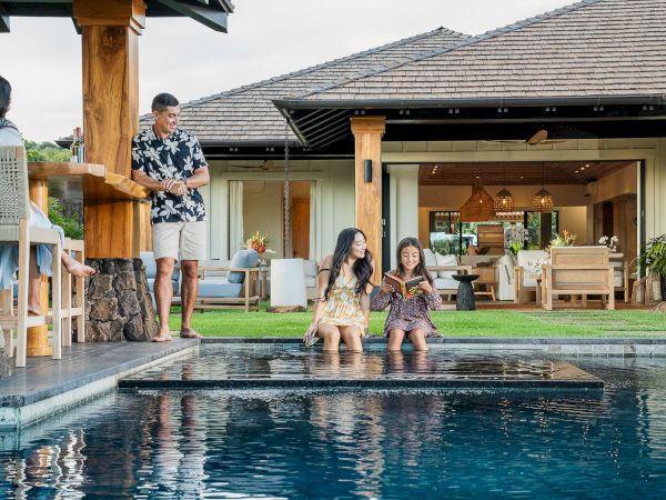 A man talks with a family by a house pool as two girls read and splash in the water.