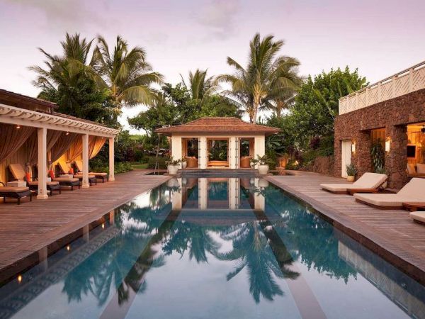 A tropical resort courtyard with a wooden deck, a swimming pool reflecting palm trees, and sun loungers under warm evening lights.