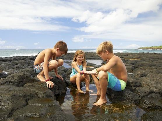Three children in swimsuits play on rocky lava shoreline, splashing and building a small sand-and-water scene by the ocean.