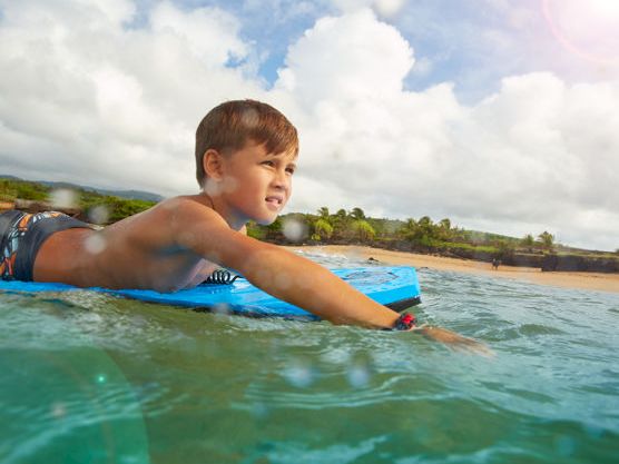 A young boy floats in ocean water on a bright blue surfboard, splashing near a sandy beach under cloudy skies.